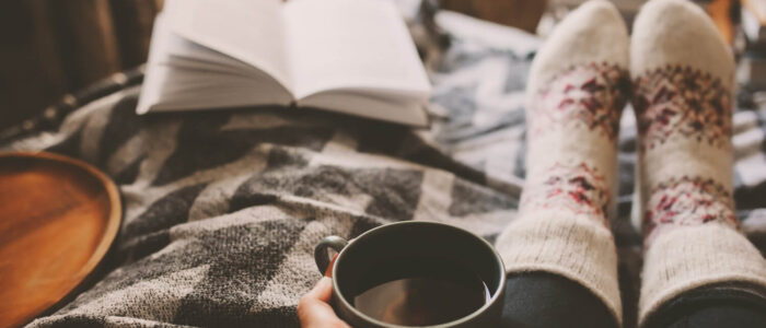 The point of view of someone looking at their feet, a cup of coffee, and a book, while embracing the spirit of Hygge.